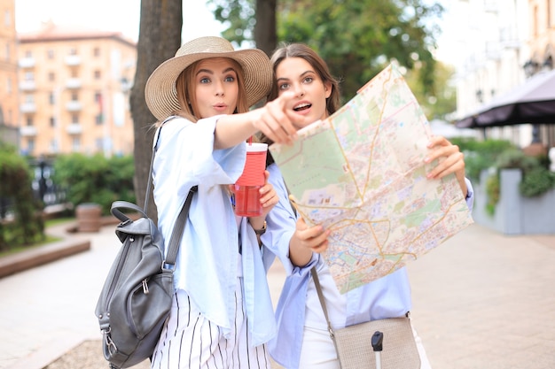 Dos chicas guapas vestidas con ropa de verano sosteniendo el mapa de la ciudad mientras están de pie al aire libre.