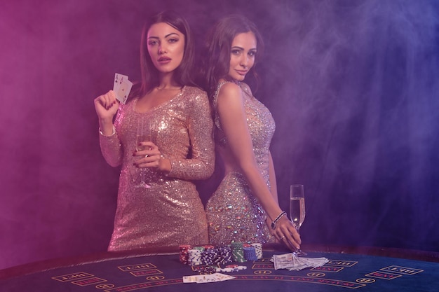 Dos chicas guapas con vestidos dorados que muestran cartas posando en la mesa de juego en el casino Fondo de humo negro luces de fondo coloridas Juegos de azar fichas de póquer dinero champán Primer plano