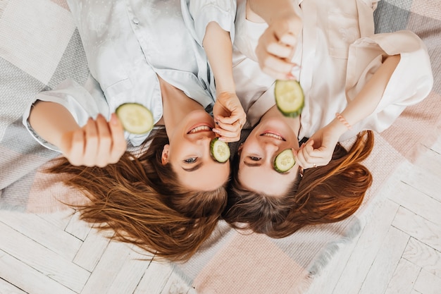 Dos chicas hacen máscaras de belleza caseras para el rostro y el cabello. Pepinos por el frescor de la piel del contorno de ojos. Las mujeres cuidan una piel joven. Las novias se ríen en casa tirado en el suelo sobre almohadas.