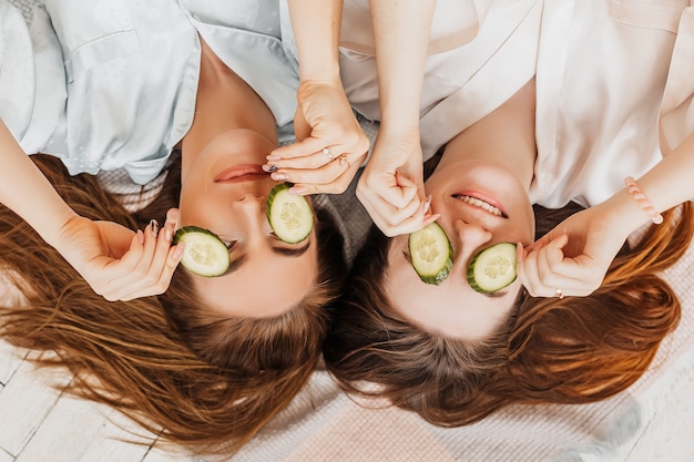 Dos chicas hacen máscaras de belleza caseras para el rostro y el cabello. Pepinos por el frescor de la piel del contorno de ojos. Las mujeres cuidan una piel joven. Las novias se ríen en casa tirado en el suelo sobre almohadas.