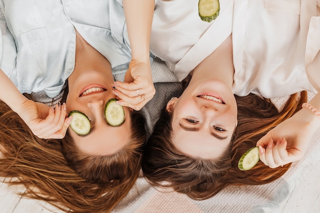 Dos chicas hacen máscaras de belleza caseras para el rostro y el cabello. Pepinos por el frescor de la piel del contorno de ojos. Las mujeres cuidan una piel joven. Las novias se ríen en casa tirado en el suelo sobre almohadas.