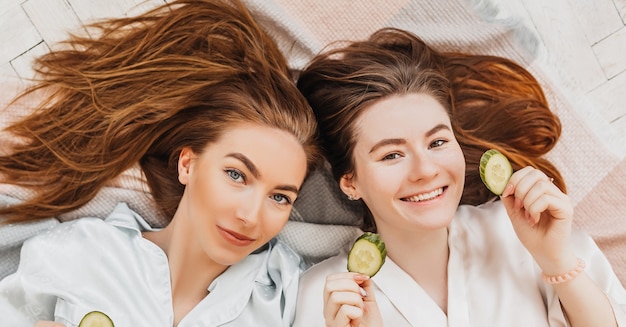 Dos chicas hacen máscaras de belleza caseras para el rostro y el cabello. Pepinos por el frescor de la piel del contorno de ojos. Las mujeres cuidan una piel joven. Las novias se ríen en casa tirado en el suelo sobre almohadas.