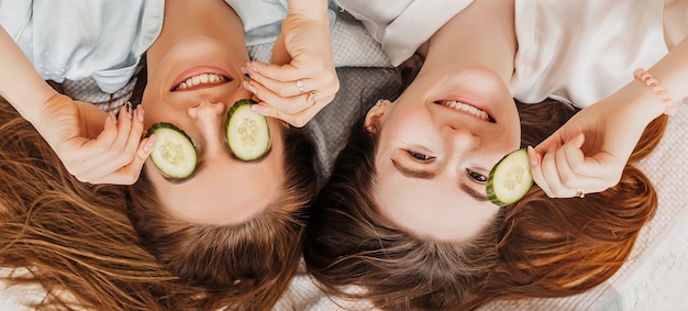 Dos chicas hacen máscaras de belleza caseras para el rostro y el cabello. Pepinos para la frescura de la piel del contorno de ojos.