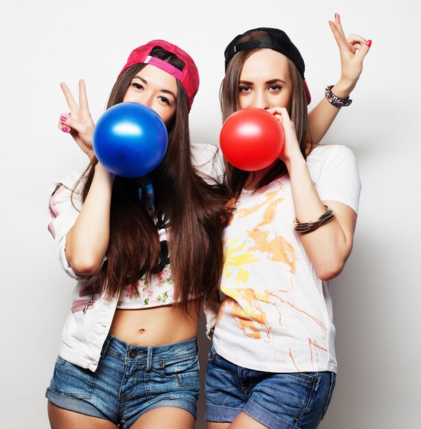 Dos chicas hipster felices sonriendo y sosteniendo globos de colores sobre fondo blanco.