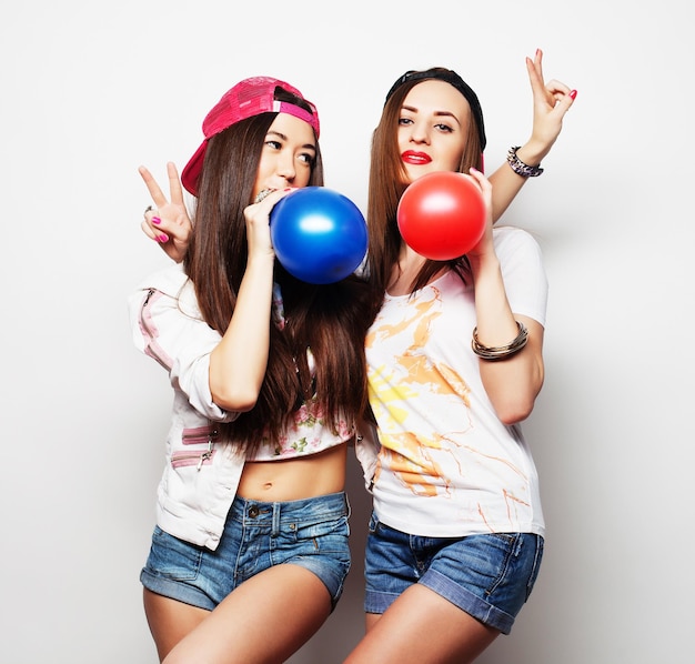 Dos chicas hipster felices sonriendo y sosteniendo globos de colores sobre fondo blanco.