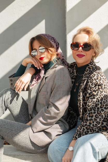 Dos chicas hipster de moda con gafas de sol vintage en ropa de moda se sientan en la calle