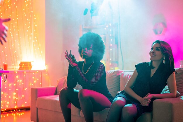 Dos chicas interculturales sentadas en el sofá mientras una de ellas habla con un amigo y la otra disfruta del descanso después de bailar en la fiesta en casa