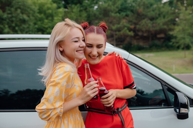 Foto dos chicas jóvenes con una botella de bebidas junto al auto