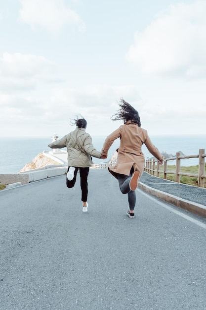 Dos chicas jóvenes corriendo juntas tomándose de la mano hacia la orilla del mar y un faro. Concepto de amistad y diversión juntos. Toma mi mano y ven conmigo concepto