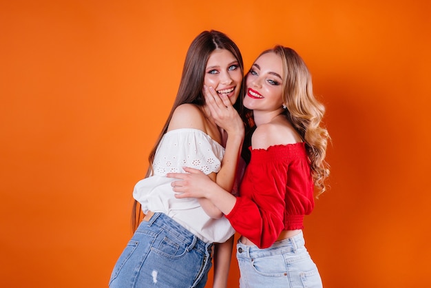 Dos chicas jóvenes y hermosas muestran emociones y sonrisas en el estudio sobre un fondo naranja. Chicas para publicidad.