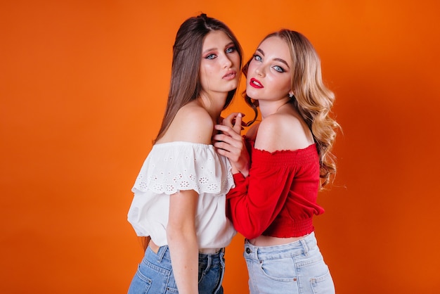 Dos chicas jóvenes y hermosas muestran emociones y sonrisas en el estudio sobre un fondo naranja. Chicas para publicidad.