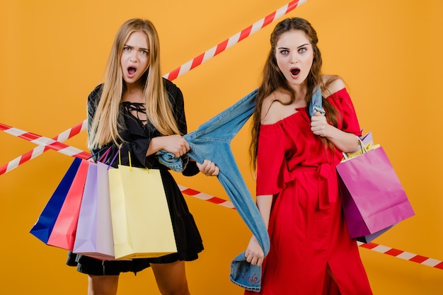 Dos chicas jóvenes y hermosas sorprendidas peleando por un par de jeans con coloridos bolsos de compras y cinta de señal aislada sobre amarillo