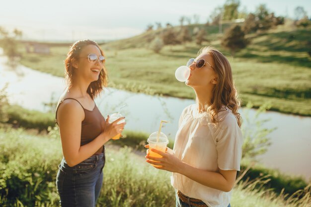 Dos chicas jóvenes infla un chicle. beber jugo del vaso de plástico, al atardecer, expresión facial positiva, al aire libre