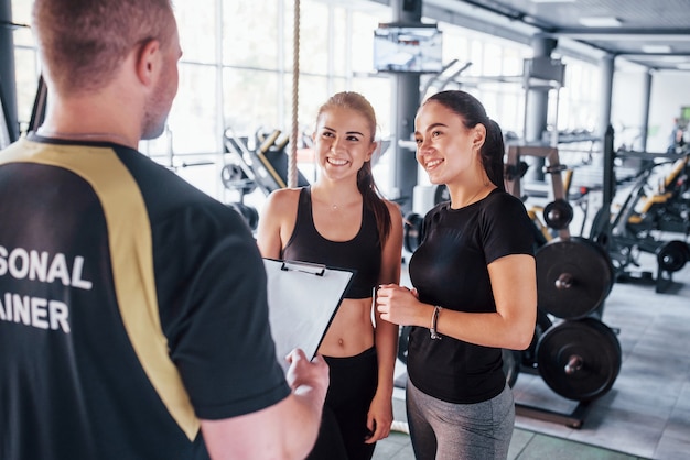 Dos chicas jóvenes en ropa deportiva está en el gimnasio junto durante el día con su entrenador personal masculino.