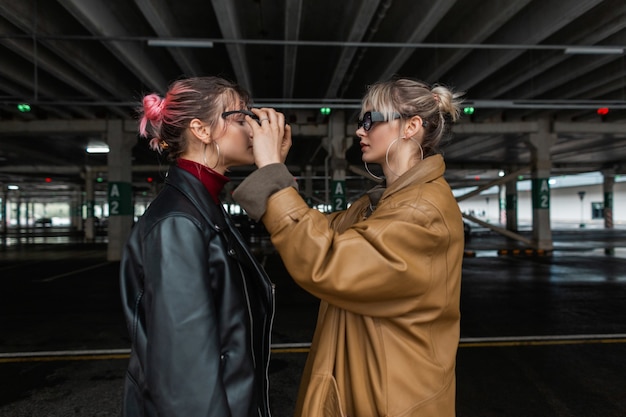 Dos chicas jóvenes con ropa de otoño de moda con chaqueta de cuero vintage con gafas de sol en la calle. Chica elegante pone gafas de sol a la chica de la ciudad
