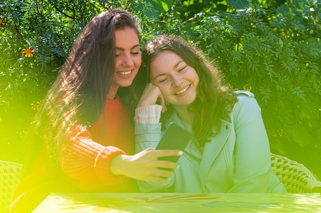 Foto dos chicas jóvenes se sientan en un café verde hablando, riendo y tomando fotos en un teléfono inteligente