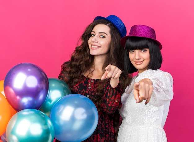 Dos chicas jóvenes sonrientes y complacidas con gorro de fiesta de pie detrás de globos mirando y apuntando aislado en la pared rosa