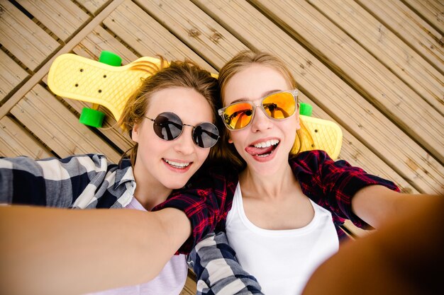 Dos chicas jóvenes en traje de hipster haciendo selfie mientras está acostado en el muelle de madera.