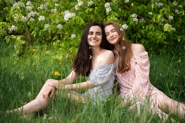 Dos chicas jóvenes vestidas están sentadas en la hierba verde debajo de un árbol blanco y riéndose