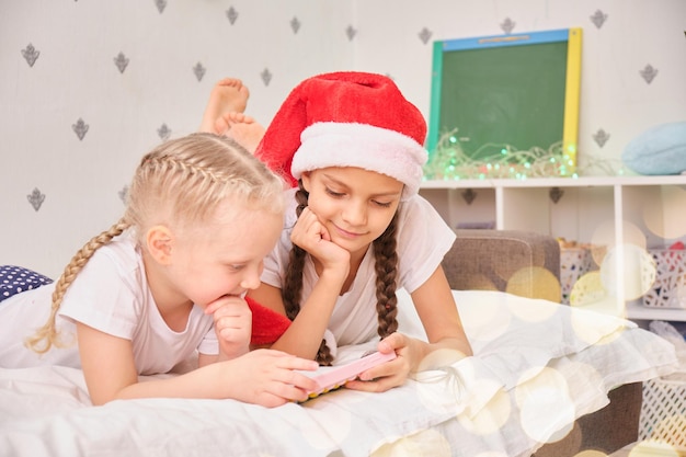 Dos chicas lindas se acuestan en la cama y juegan juegos de teléfonos inteligentes, concepto de vacaciones navideñas, videollamadas a familiares para feliz navidad, decoración festiva y estado de ánimo