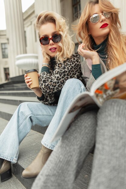 Dos chicas de moda con ropa elegante sentadas en los escalones tomando café y leyendo una revista en la ciudad