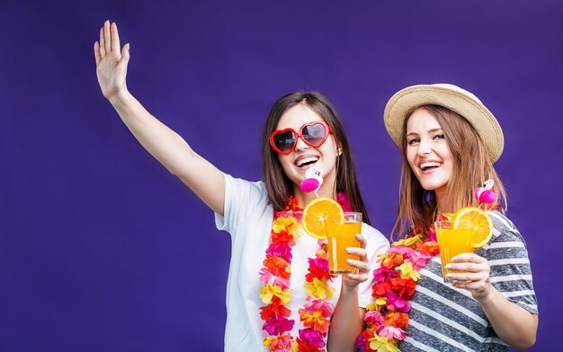 Dos chicas muy sonrientes con lei en el cuello sostienen bebidas naranjas antes de un fondo morado