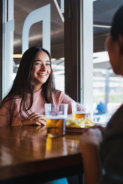 Dos chicas sonrientes hablando entre ellas en un bar. Están tomando unas cervezas y un bocadillo.