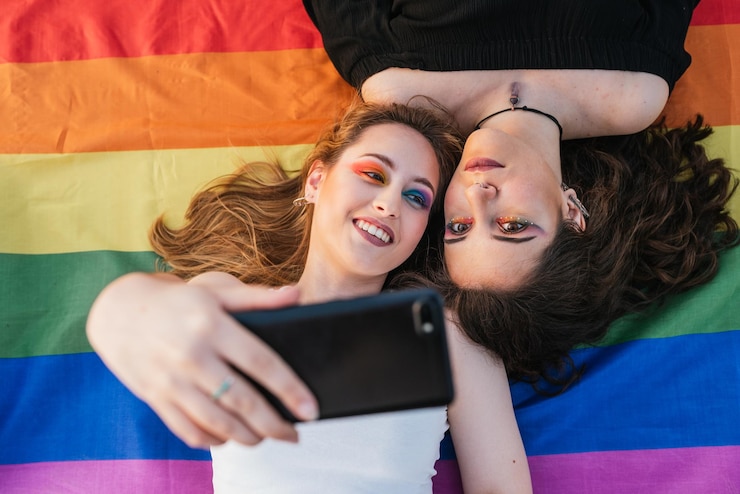 Foto dos chicas tomándose una foto con sus teléfonos móviles sobre un fondo colorido lgtb