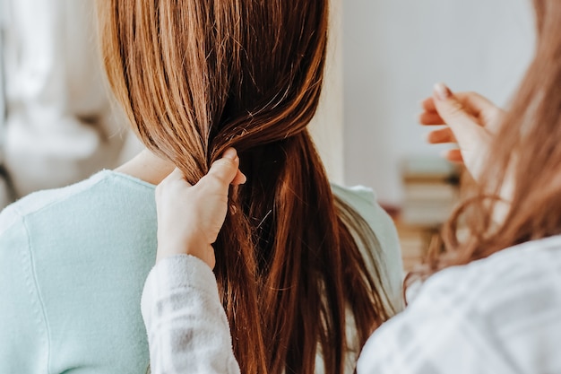 Dos chicas se trenzan el pelo en la ventana. La mujer le hace una trenza a su amiga. Peinados para tejer el cabello. Novia trenza sus manos con rizos. Cuidado del cabello