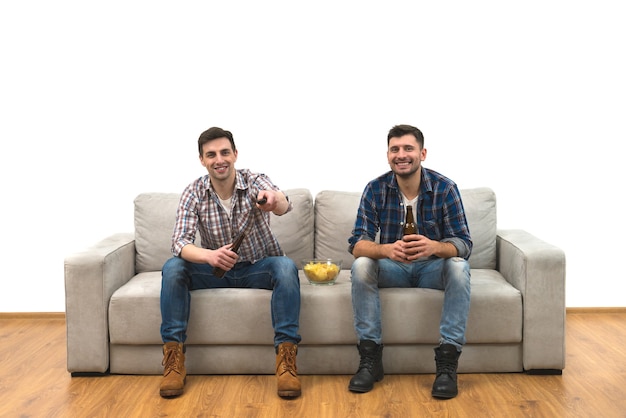 Los dos chicos beben una cerveza con patatas fritas en el sofá sobre una pared blanca de fondo