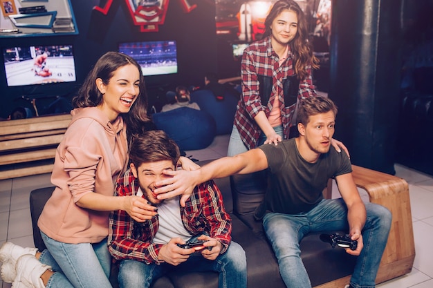 Dos chicos jugando en el sofá en la habitación. Uno de ellos toma la mano frente a la cara del oponente. Mujeres jóvenes riendo. Están detrás de los hombres.