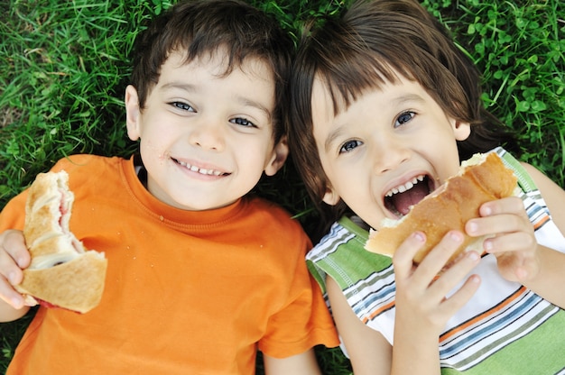 Dos chicos lindos tirado en el suelo en la naturaleza y felizmente comiendo alimentos saludables