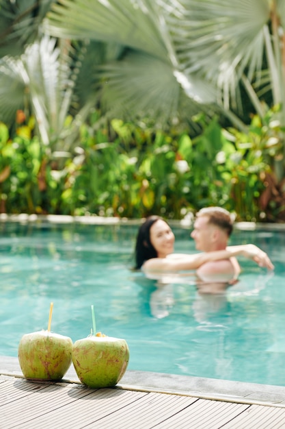 Dos cócteles de coco en el borde de la piscina, pareja bailando y abrazándose en el agua