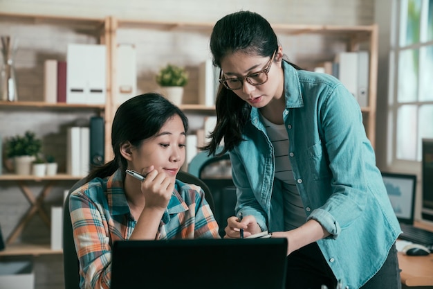 dos colegas asiáticas del espacio de coworking de trabajo moderno hablando y sonriendo mientras se sientan en la mesa del escritorio con el bolígrafo de la computadora portátil apuntando en el documento del cuaderno. jovencita nueva fresca preguntar amigable gerente