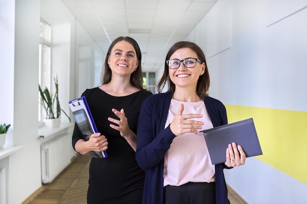 Dos colegas de la empresaria, maestra caminando y hablando en el pasillo de la oficina, la escuela. Mujeres jóvenes y de mediana edad sonrientes positivas, concepto de trabajadores de oficina de profesiones de educación empresarial