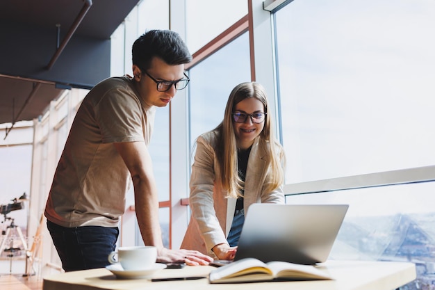 Dos colegas, un hombre y una mujer gerente, se paran cerca de una mesa con una computadora portátil en una oficina moderna y discuten un nuevo proyecto Dos socios comerciales con anteojos están hablando de un nuevo negocio