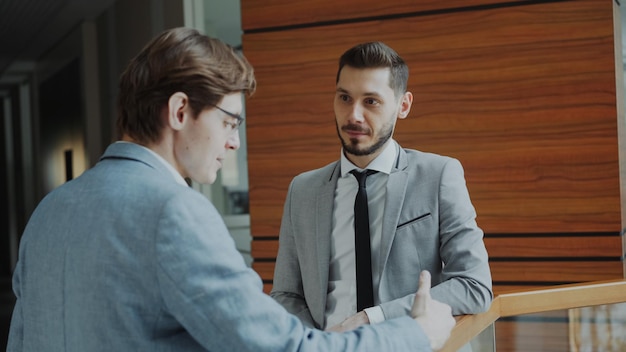 Dos colegas de negocios de pie y charlando de pie cerca de la baranda en el pasillo