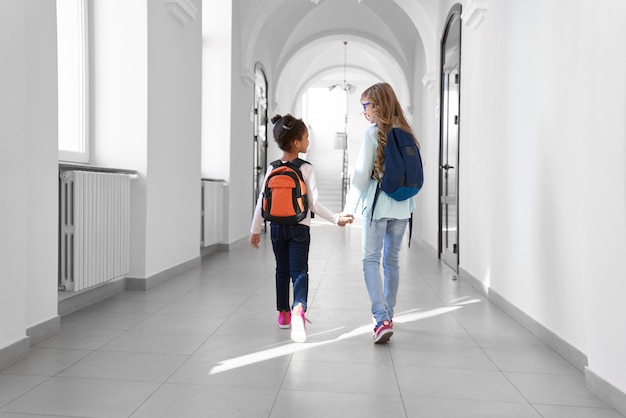 Dos colegialas con vaqueros y zapatillas de deporte con mochilas caminando en un largo y ligero pasillo después de las clases.