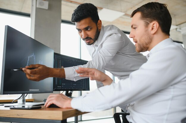 Dos comerciantes exitosos en ropa formal apuntando a la pantalla analizando estadísticas y dinámicas en gráficos forex trabajando juntos en la oficina