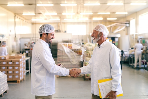 Dos compañeros de trabajo felices dándose la mano mientras está de pie en la fábrica de alimentos.