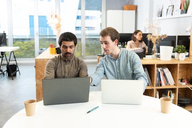 Dos compañeros de trabajo masculinos que trabajan en sus computadoras portátiles ayudándose unos a otros.