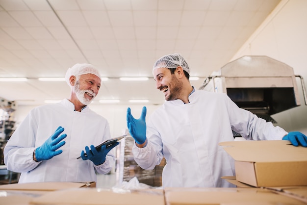 Dos compañeros varones i ropa estéril preparando cajas con productos para su transporte. De pie en una habitación luminosa o un almacén trabajando juntos y riendo.