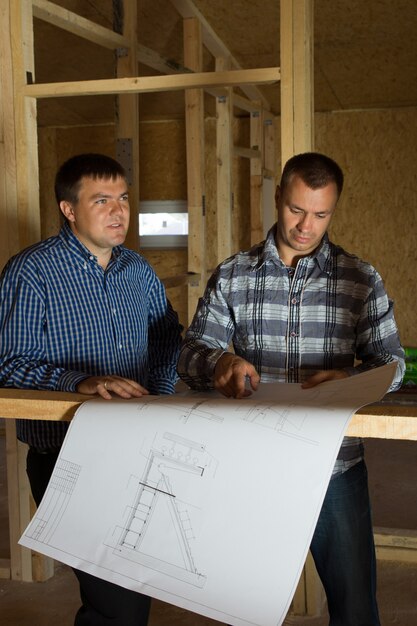 Dos constructores comprobando un plano juntos mientras están parados dentro de una casa de estructura de madera a medio construir