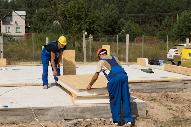 Dos constructores o trabajadores de la construcción que manipulan paneles de pared aislados mientras se preparan para instalarlos en el piso y los cimientos de una casa de nueva construcción.