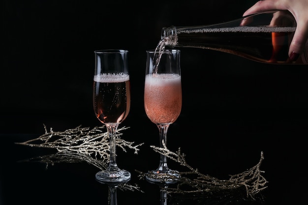 Dos copas de champán rosa y decoración de Navidad o año nuevo sobre fondo negro. Mano de mujer sostiene una botella y champán vertido. Cena romántica. Concepto de vacaciones de invierno.