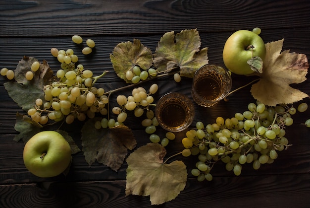 Dos copas de cristal con vino blanco, manzanas, uvas y hojas secas sobre una superficie de madera. Vista superior