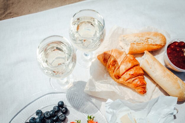 Dos copas de vino blanco y bayas, pasteles, pan y queso camembert en mesa blanca configurada