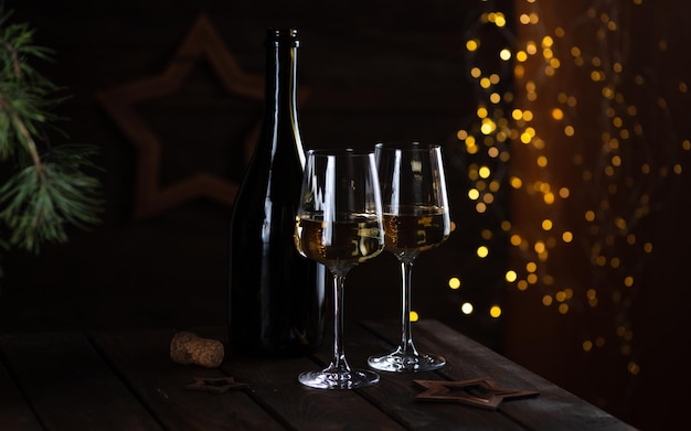 Dos copas de vino blanco y una botella sobre una mesa de madera y guirnaldas de luces detrás