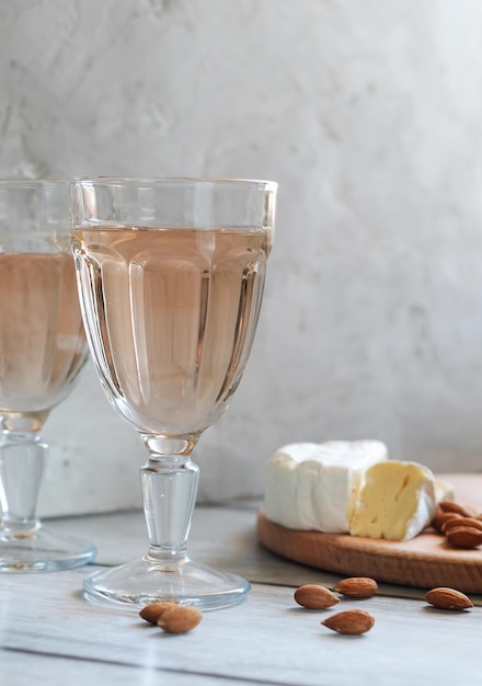 Dos copas de vino rosado junto a queso brie y almendras en una tabla de cortar de madera. Fondo de hormigón ligero Orientación vertical.