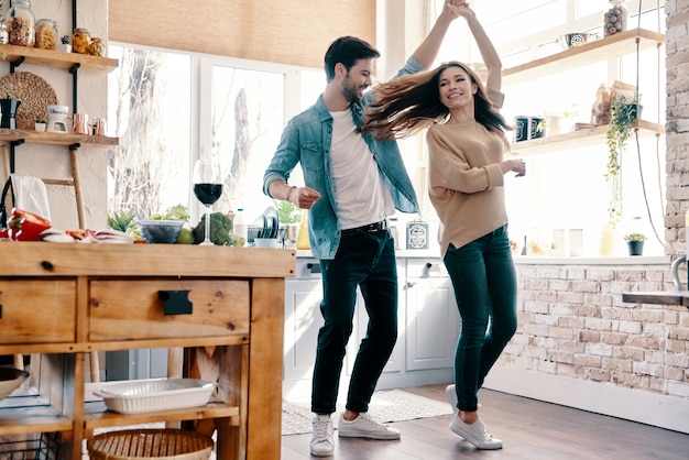 Dos corazones llenos de amor. Longitud total de la hermosa joven pareja en ropa casual bailando y sonriendo mientras está de pie en la cocina de casa